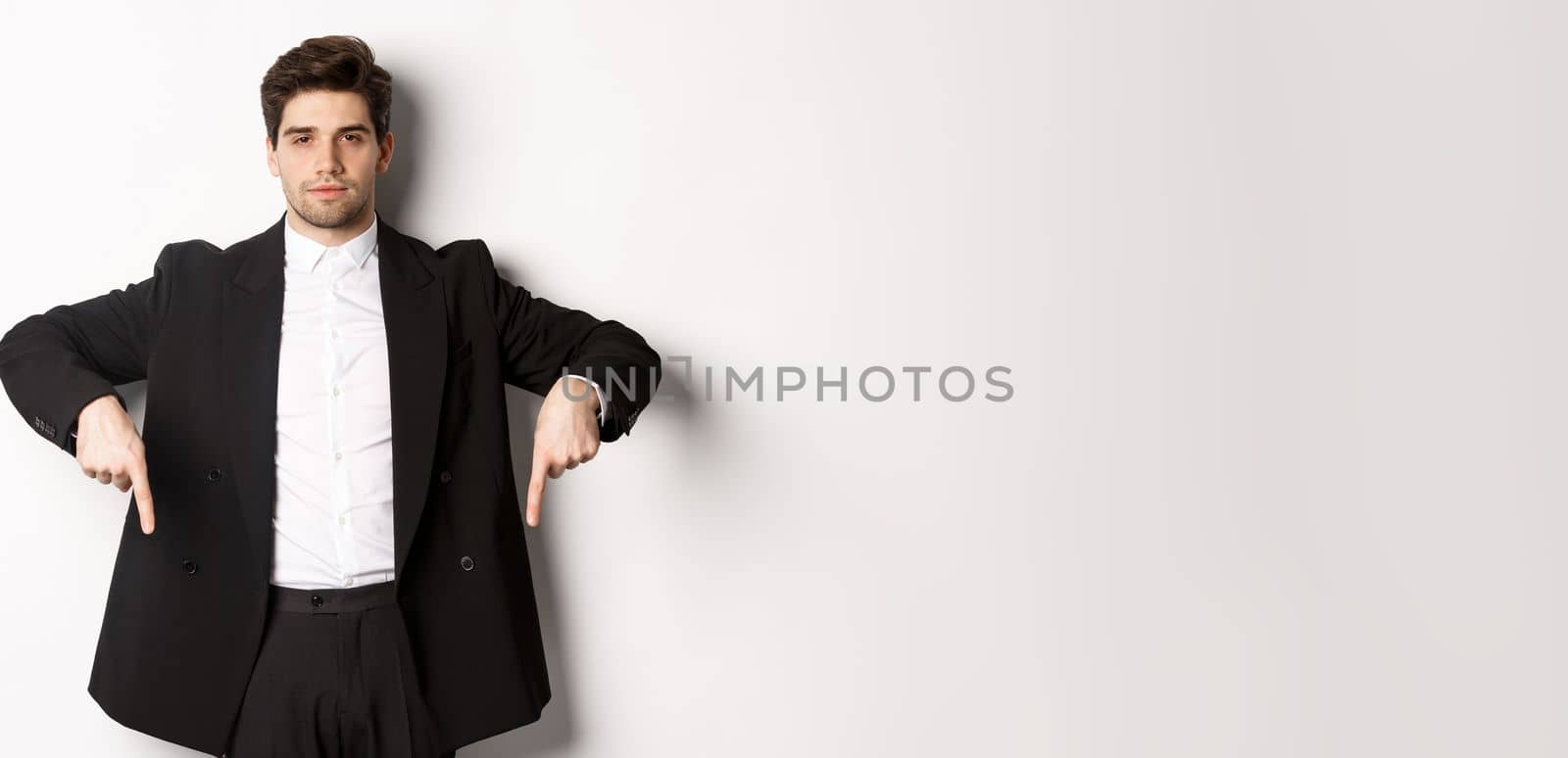 Image of handsome man dressed for formal party, wearing suit and pointing fingers down, showing advertisement or making announcement, standing over white background.