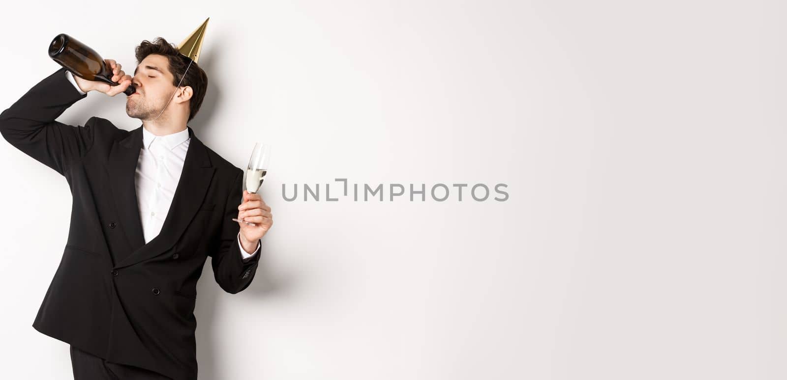 Image of handsome guy in trendy suit, drinking champagne from bottle and celebrating holiday, having a birthday party, getting drunk while standing over white background.