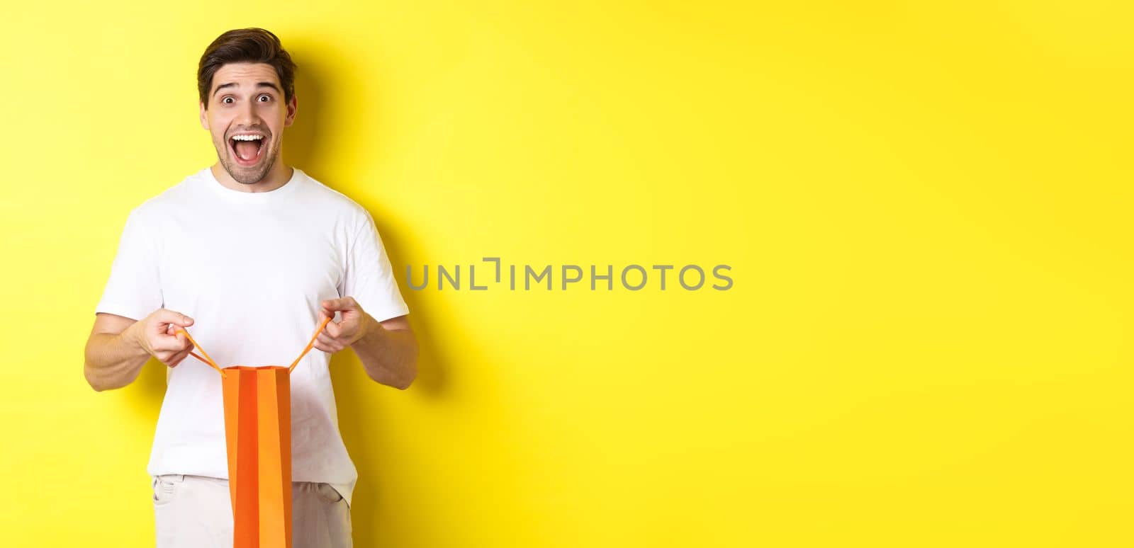 Surprised guy open shopping bag with fist, looking excited and happy at camera, standing against yellow background by Benzoix