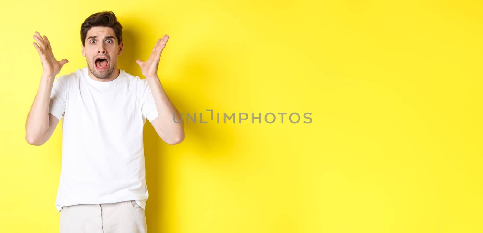 Frustrated and shocked guy panicking, screaming and looking scared, standing in white t-shirt over yellow background.