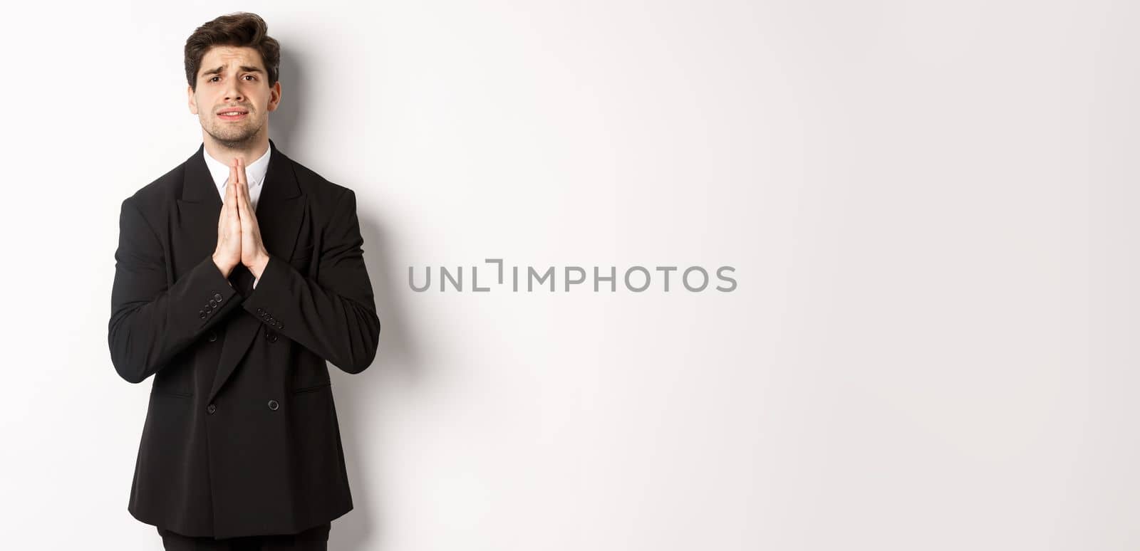 Image of handsome businessman in suit need help, begging you, holding hands in plead gesture and looking desperate, standing over white background.