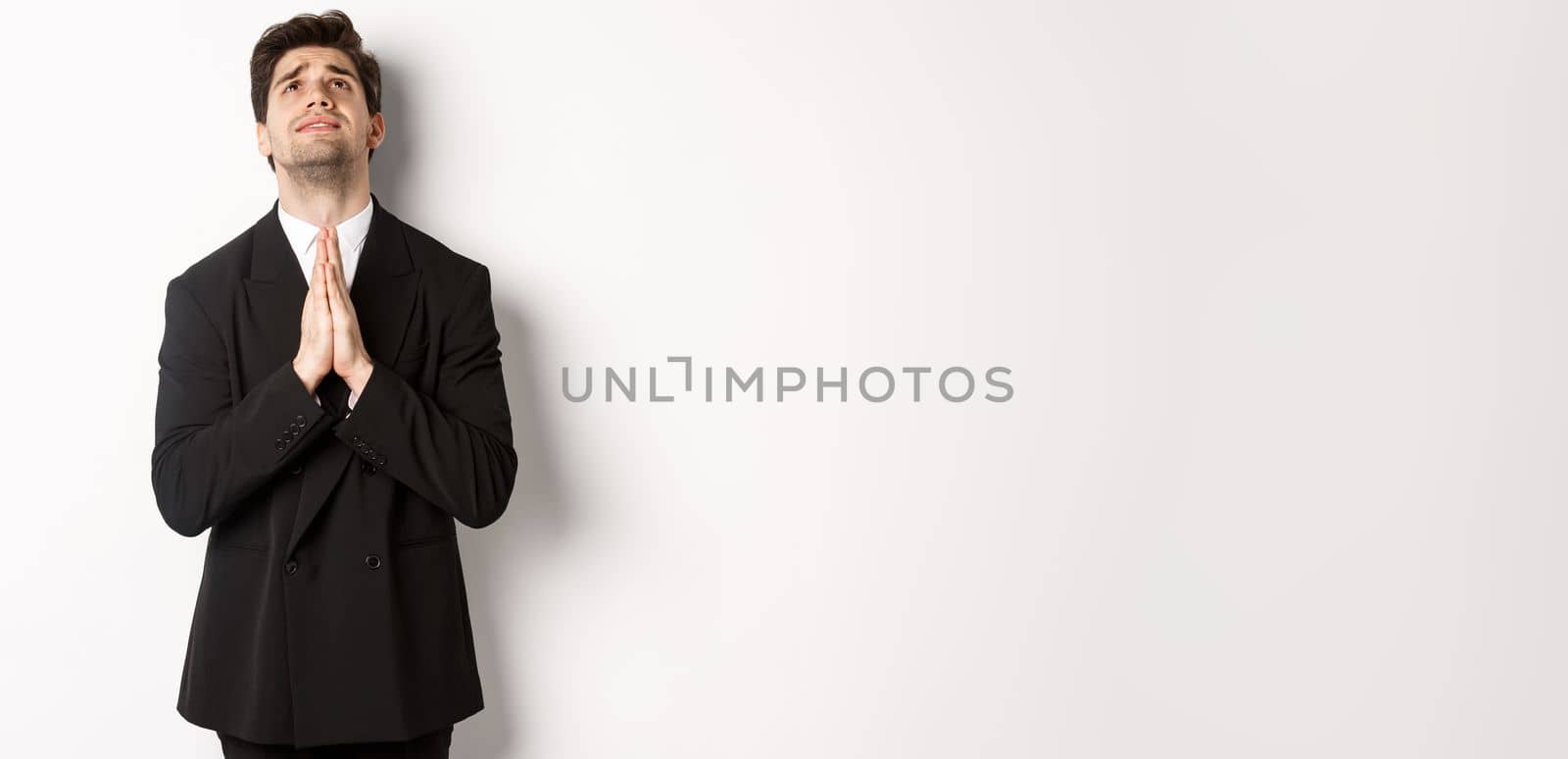 Troubled and hopeful man in black suit begging god, pleading and looking up, need help, standing over white background.