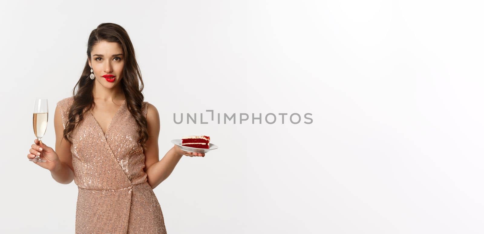 Party and celebration concept. Sexy woman in elegant dress, holding champagne and piece of cake, biting lip and looking seductive at camera.