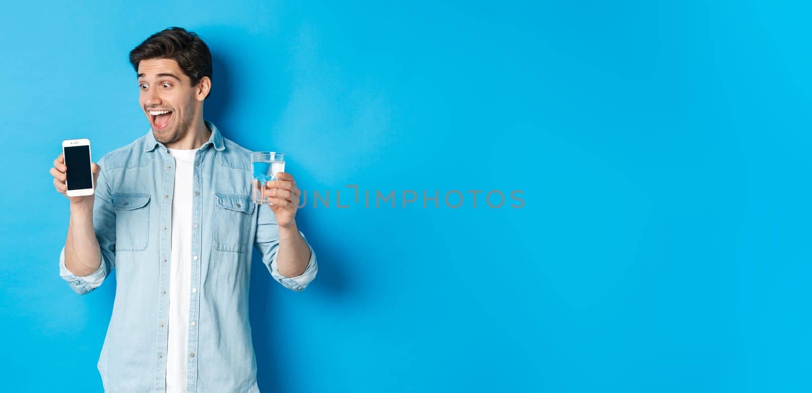 Happy man looking excited at mobile phone screen, holding glass of water, standing over blue background.