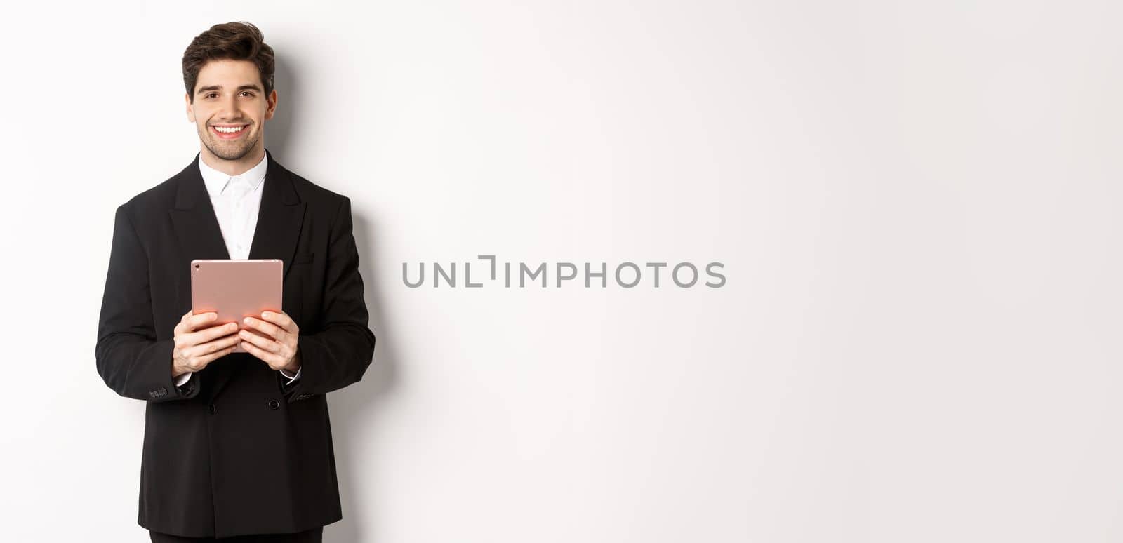 Image of handsome businessman in trendy suit, holding digital tablet and smiling, standing against white background.