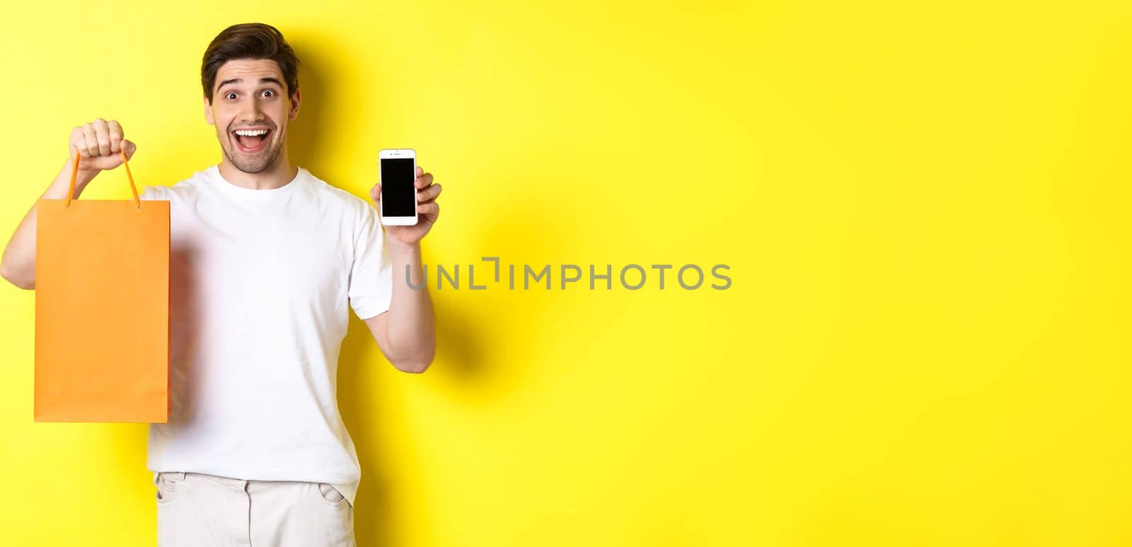 Surprised man holding shopping bag and showing smartphone screen, concept of mobile banking and app achievements, yellow background.
