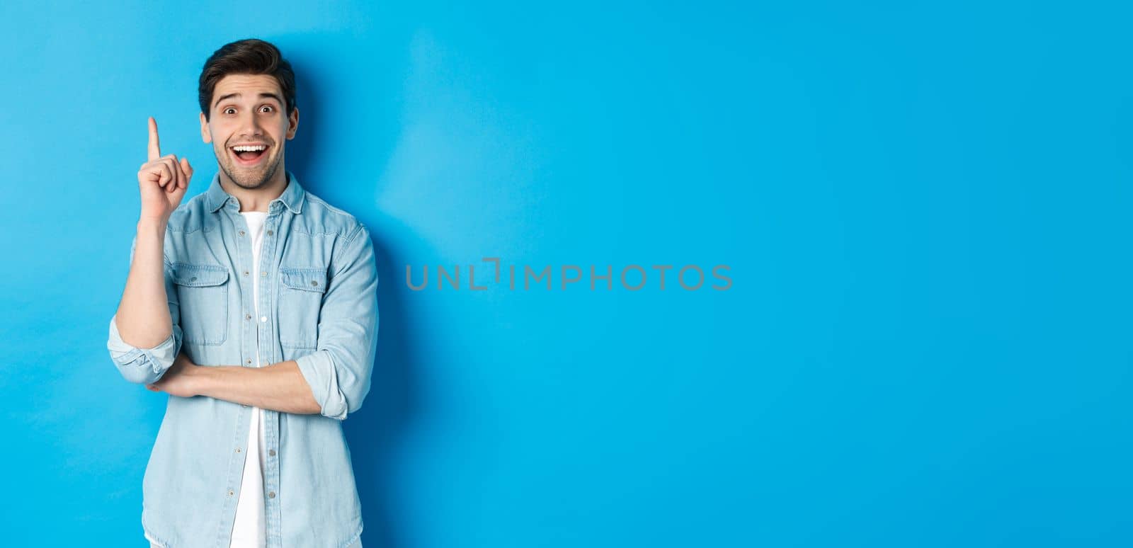 Image of handsome adult man having an idea, raising finger and smiling excited, found solution, standing against blue background.