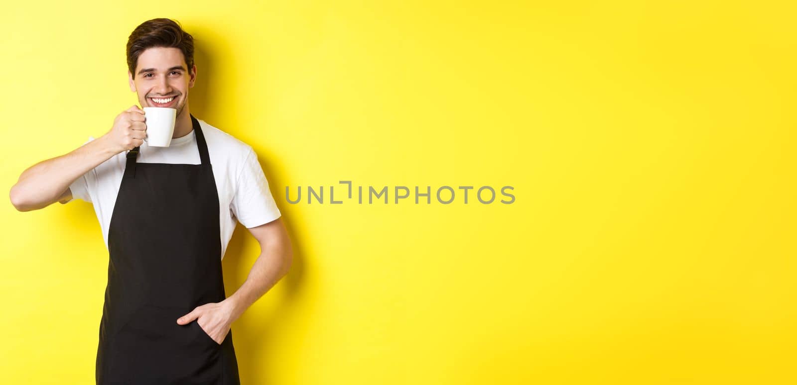 Cafe barista drinking cup of coffee and smiling, wearing black apron, standing over yellow background.