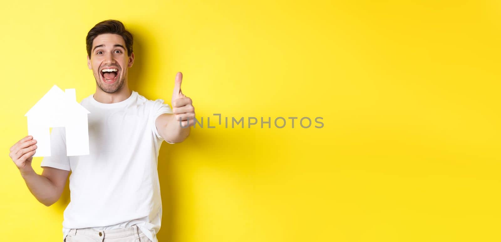 Real estate concept. Happy young male buyer showing thumb up and paper house model, smiling satisfied, standing over yellow background.