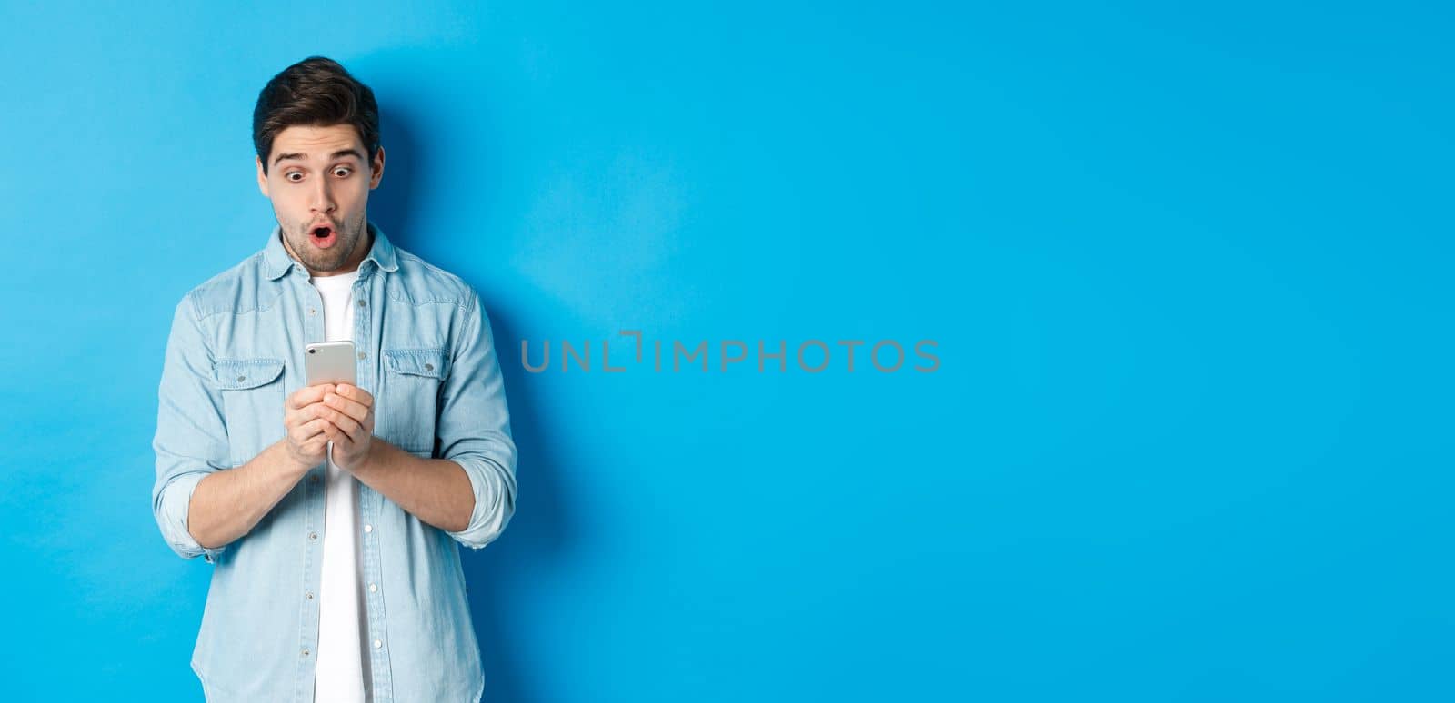 Man looking amazed while checking promo on smartphone, looking surprised at phone, standing against blue background.