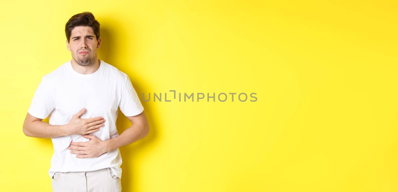 Man having stomach ache, grimacing from pain and touching belly, standing over yellow background.