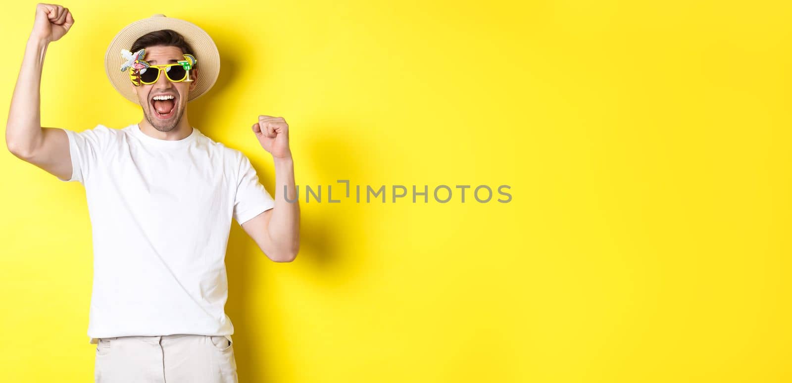 Concept of tourism and lifestyle. Happy man winning trip to resort, shouting yes and raising hands up, triumphing, wearing sunglasses and summer hat, yellow background.