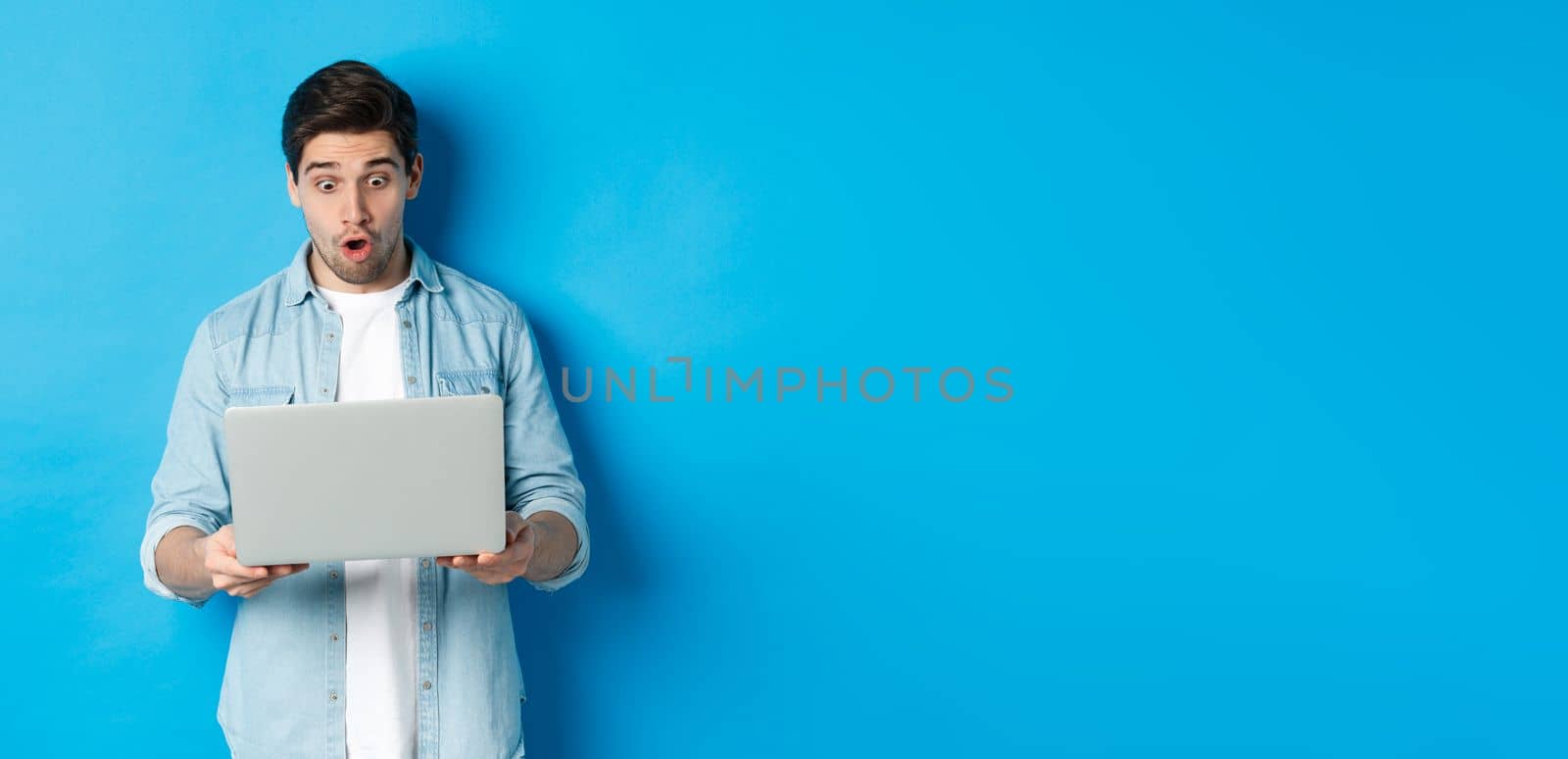 Impressed caucasian guy looking at laptop screen with amazement, checking out promo in internet, standing against blue background.