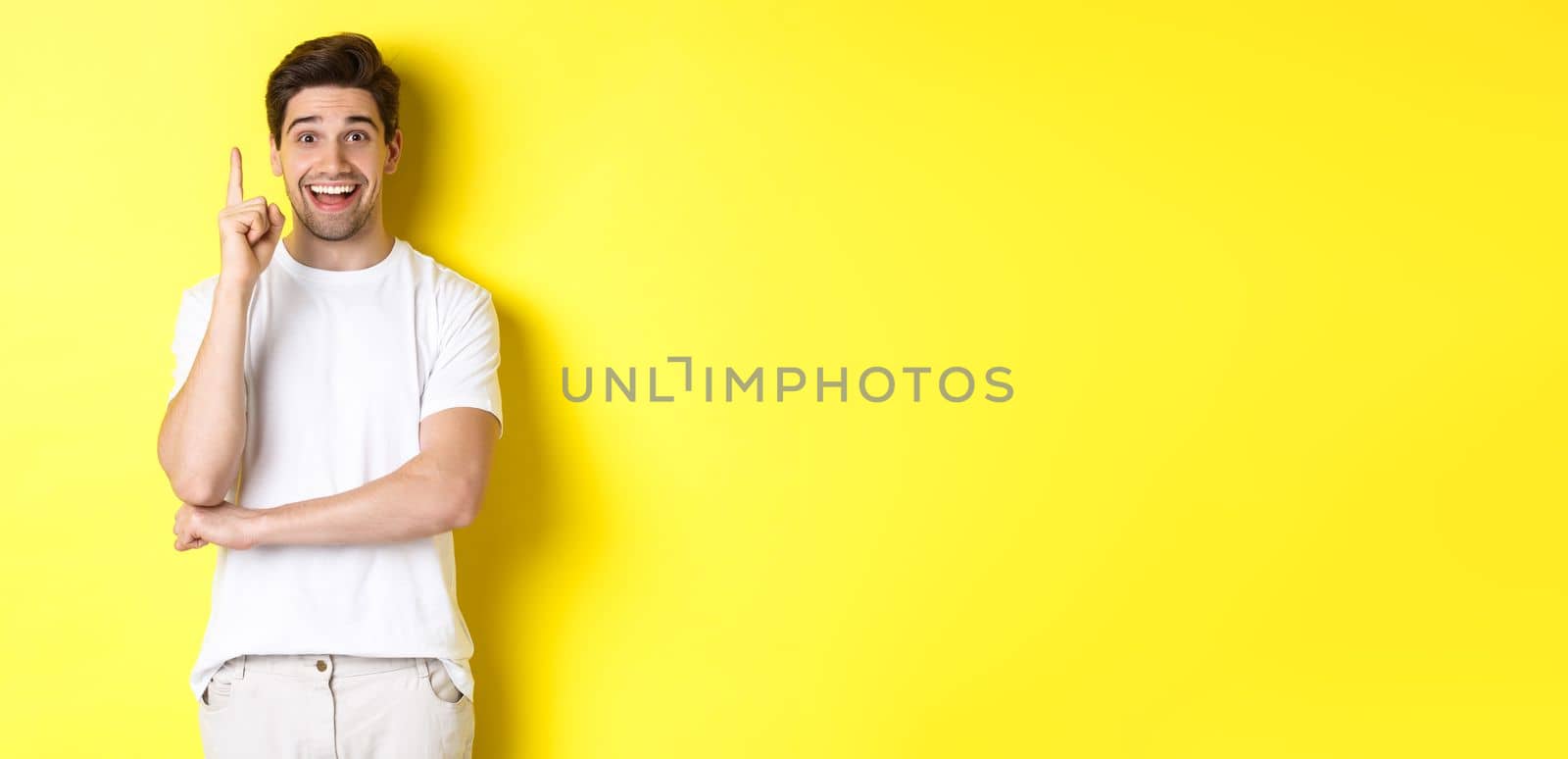 Image of attractive guy having an idea, raising finger up and suggesting plan, smiling excited, standing over yellow background.