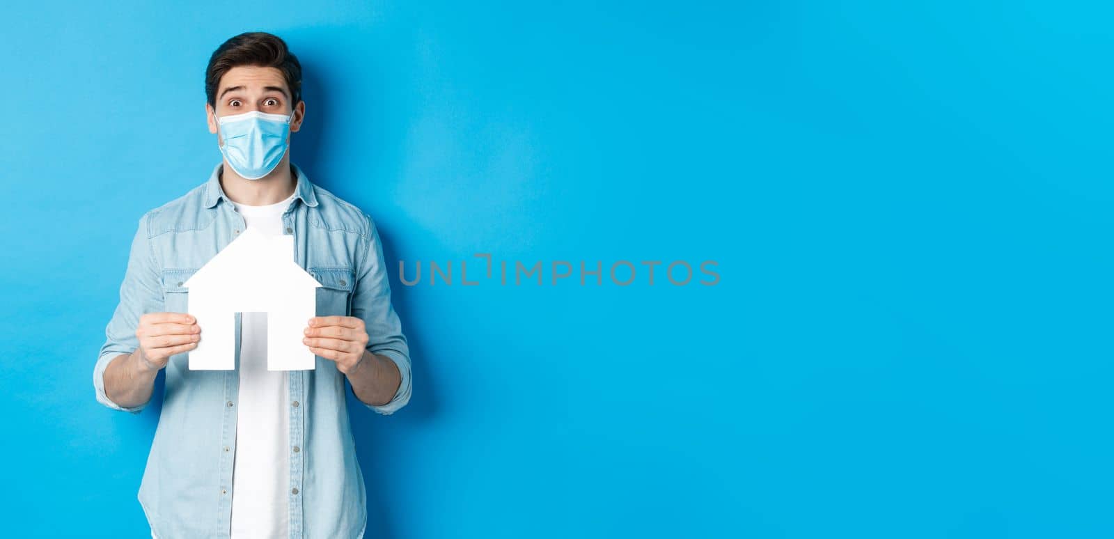 Concept of coronavirus, quarantine and social distancing. Young man searching apartment, showing house paper model, wearing medical mask, renting or buying propery, blue background.