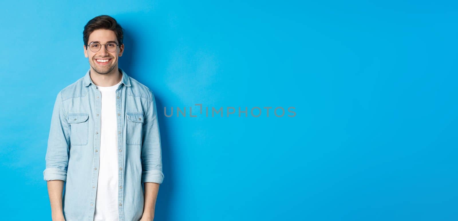 Young modern man in glasses and casual outfit standing against blue background, smiling happy at camera.