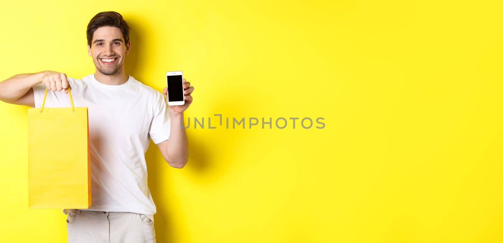 Concept of mobile banking and cashback. Young happy guy holding shopping bag and showing smartphone screen, yellow background.