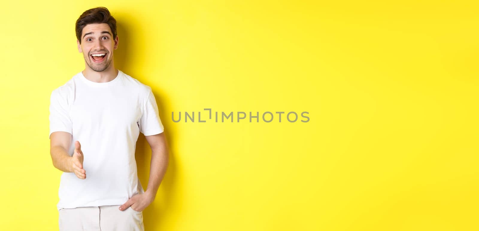 Friendly man greeting you with handshake, smiling amused, saying hello, standing over yellow background by Benzoix