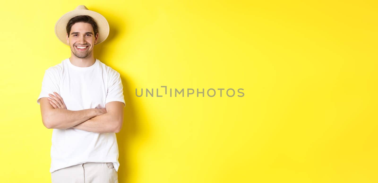 Tourism. Handsome young man looking happy, wearing straw hat for travelling, cross hands on chest and smiling, standing over yellow background.