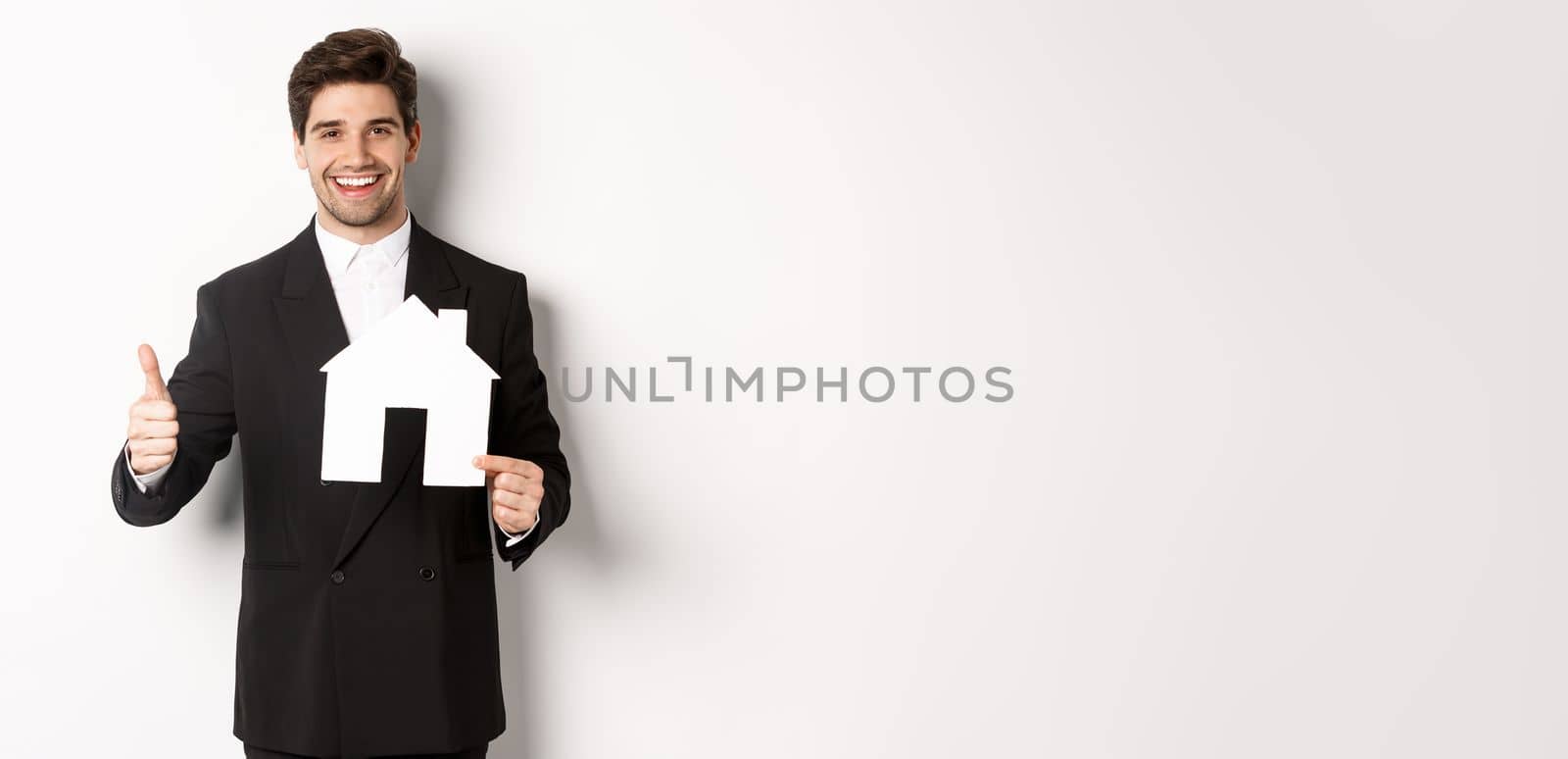 Portrait of confident real estate agent showing house maket and thumb-up, standing against white background.