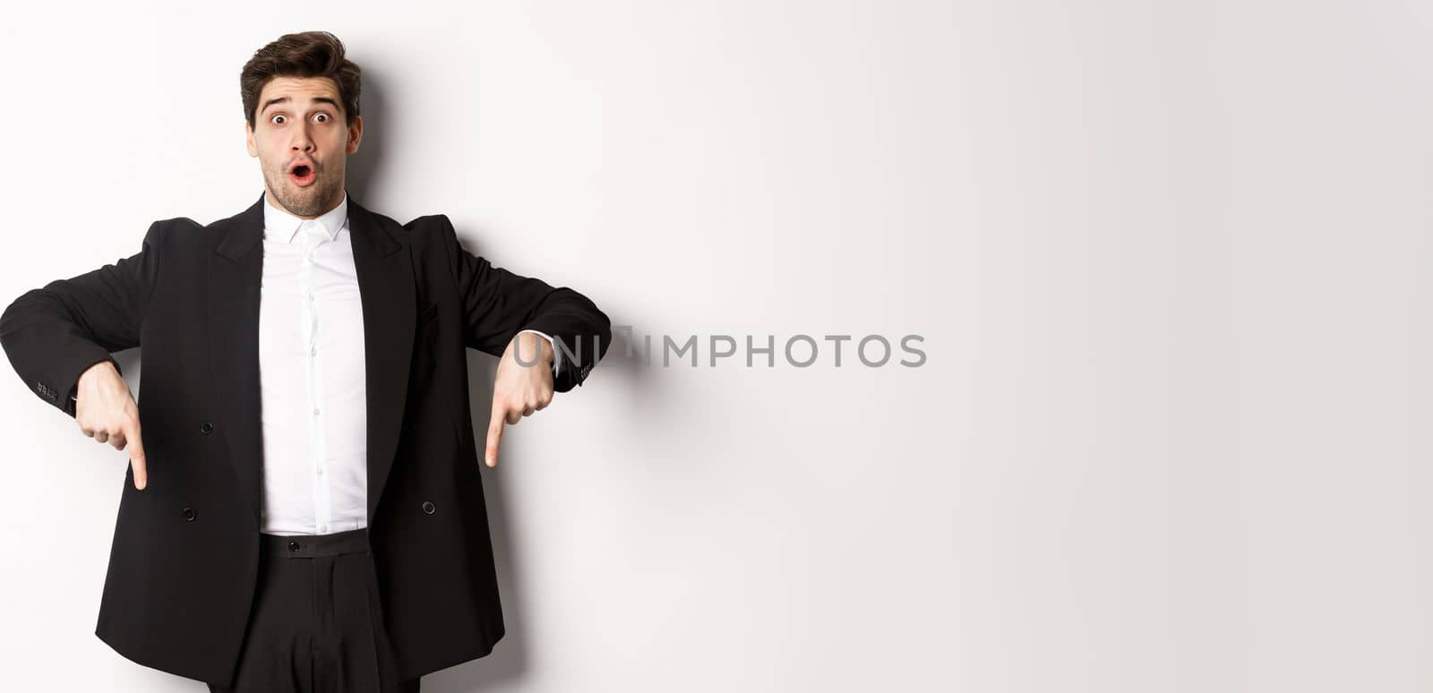 Portrait of surprised and excited man in black suit, pointing fingers down and showing christmas advertisement, standing over white background.