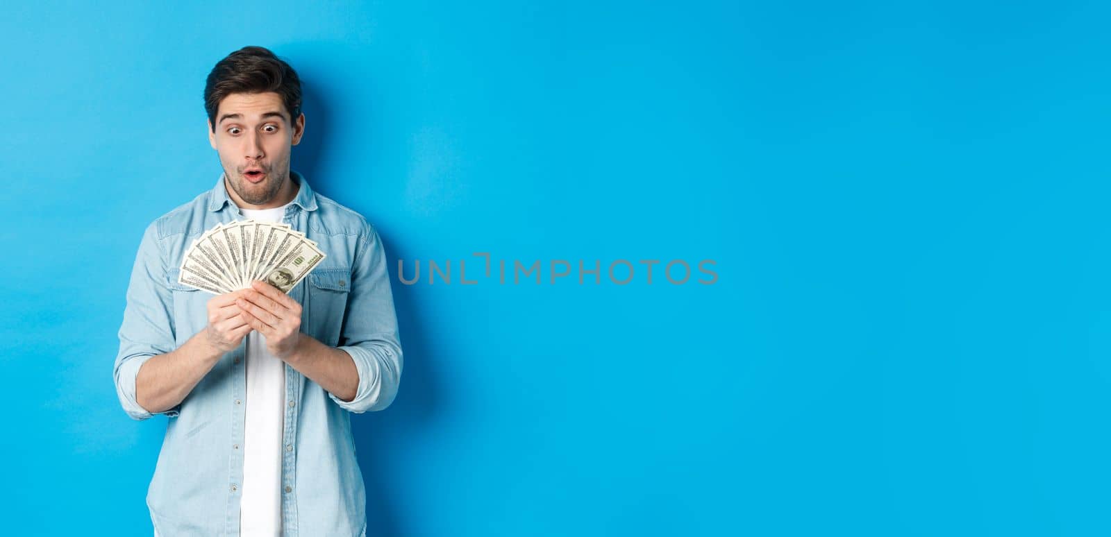 Excited successful man counting money, looking satisfied at cash and smiling, standing over blue background.