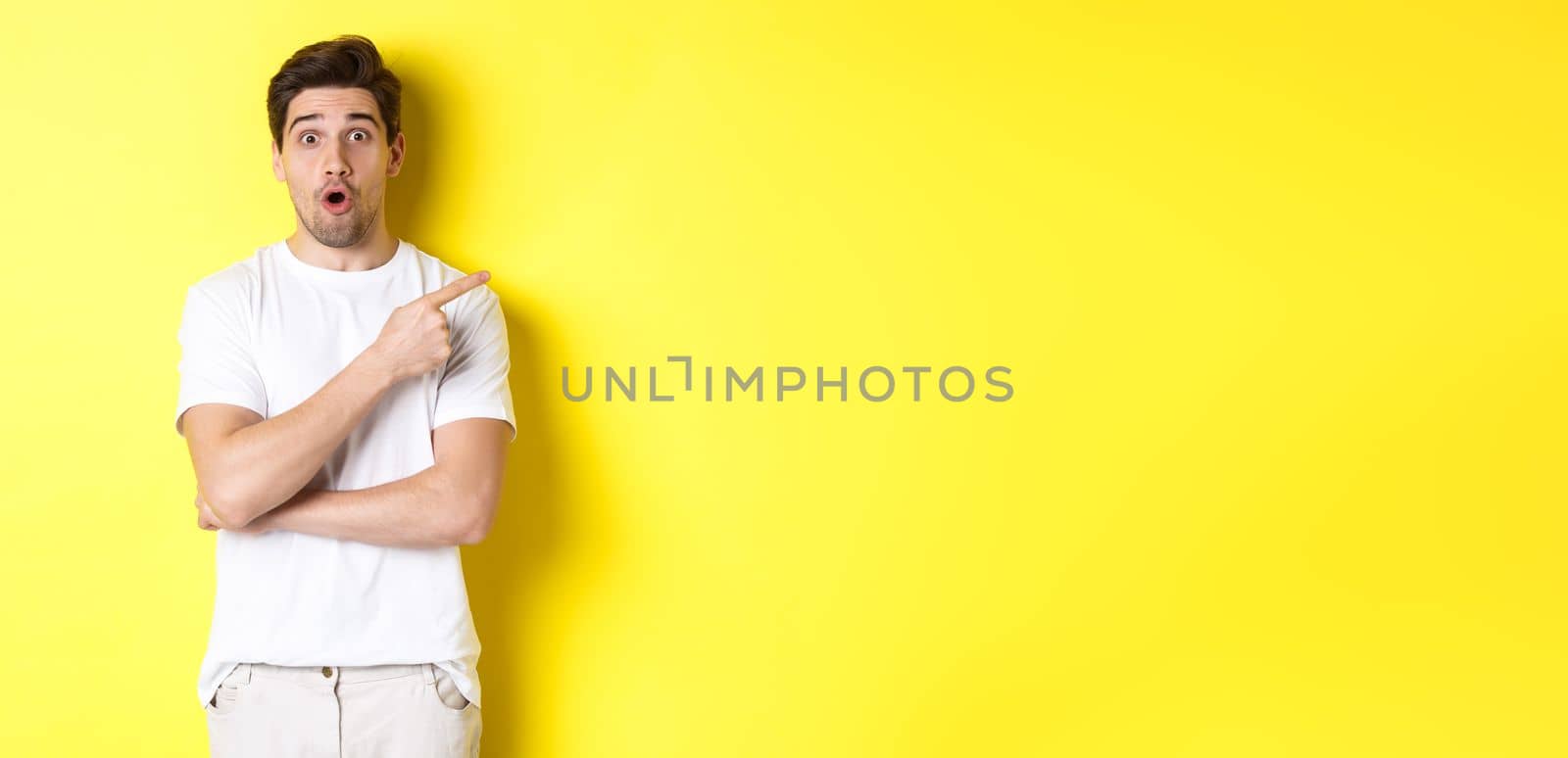 Portrait of surprised handsome man pointing finger right, gasping amazed, standing over yellow background.