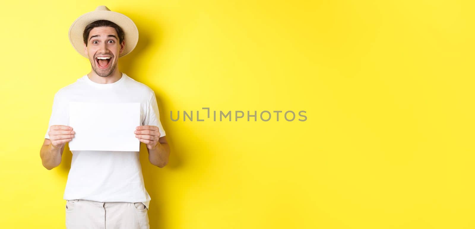 Excited tourist showing your logo or sign on blank piece of paper, smiling amazed, standing against yellow background.