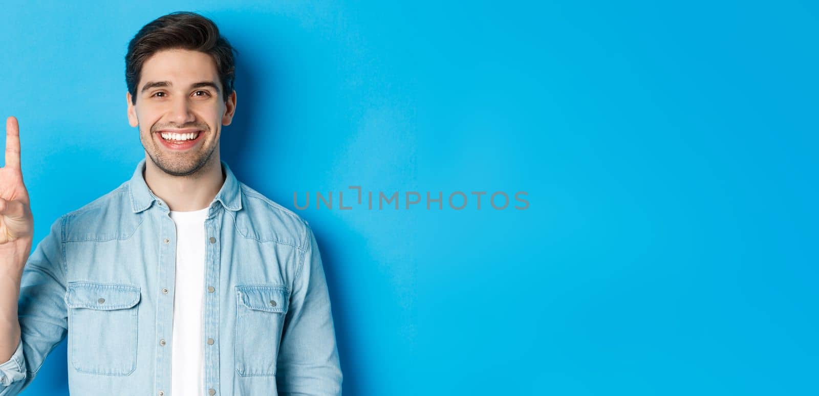 Close-up of handsome man smiling, showing fingers number three, standing over blue background by Benzoix