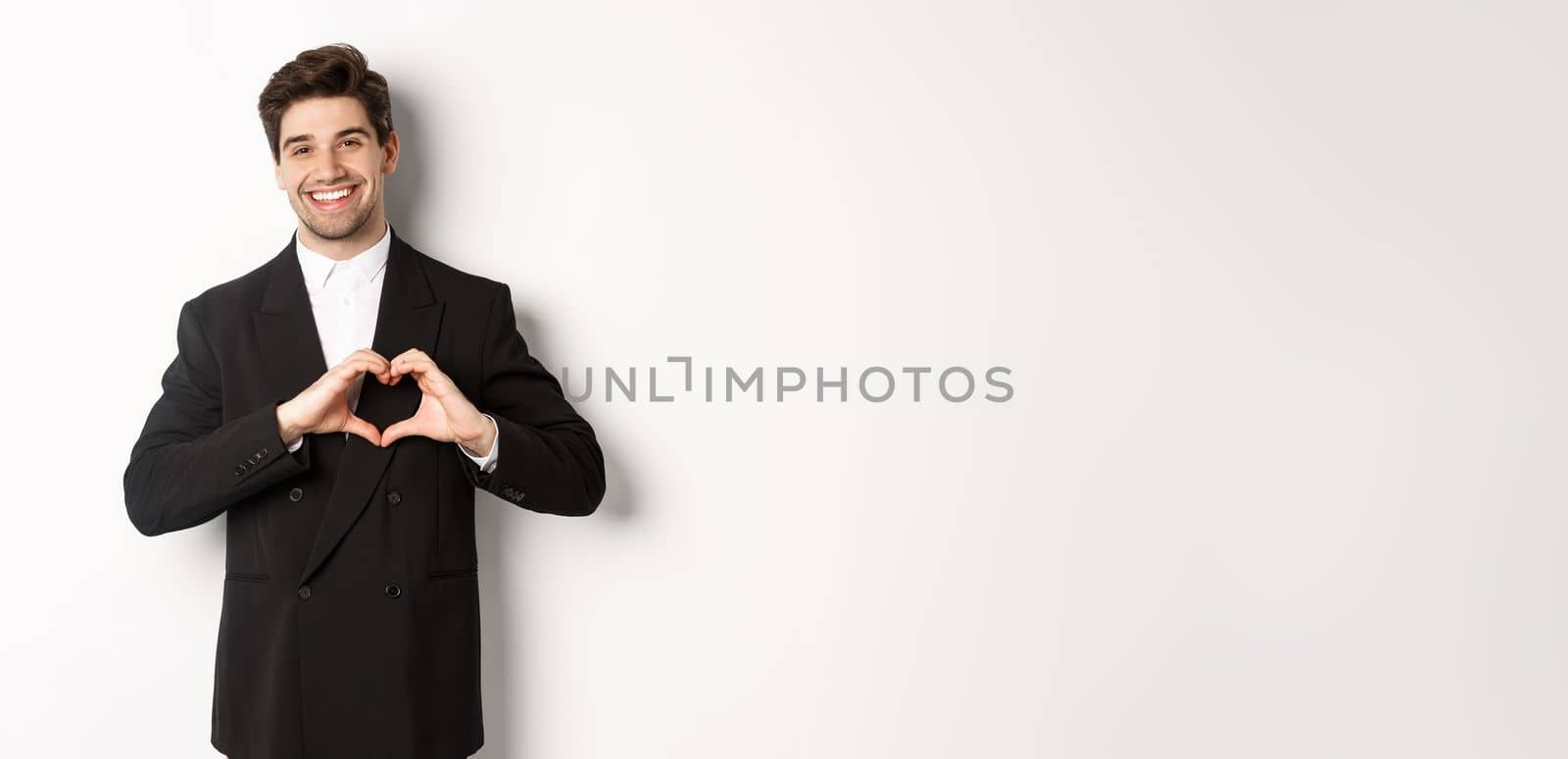 Image of handsome groom in black suit, showing heart sign and smiling, express love and sympathy, standing over white background by Benzoix