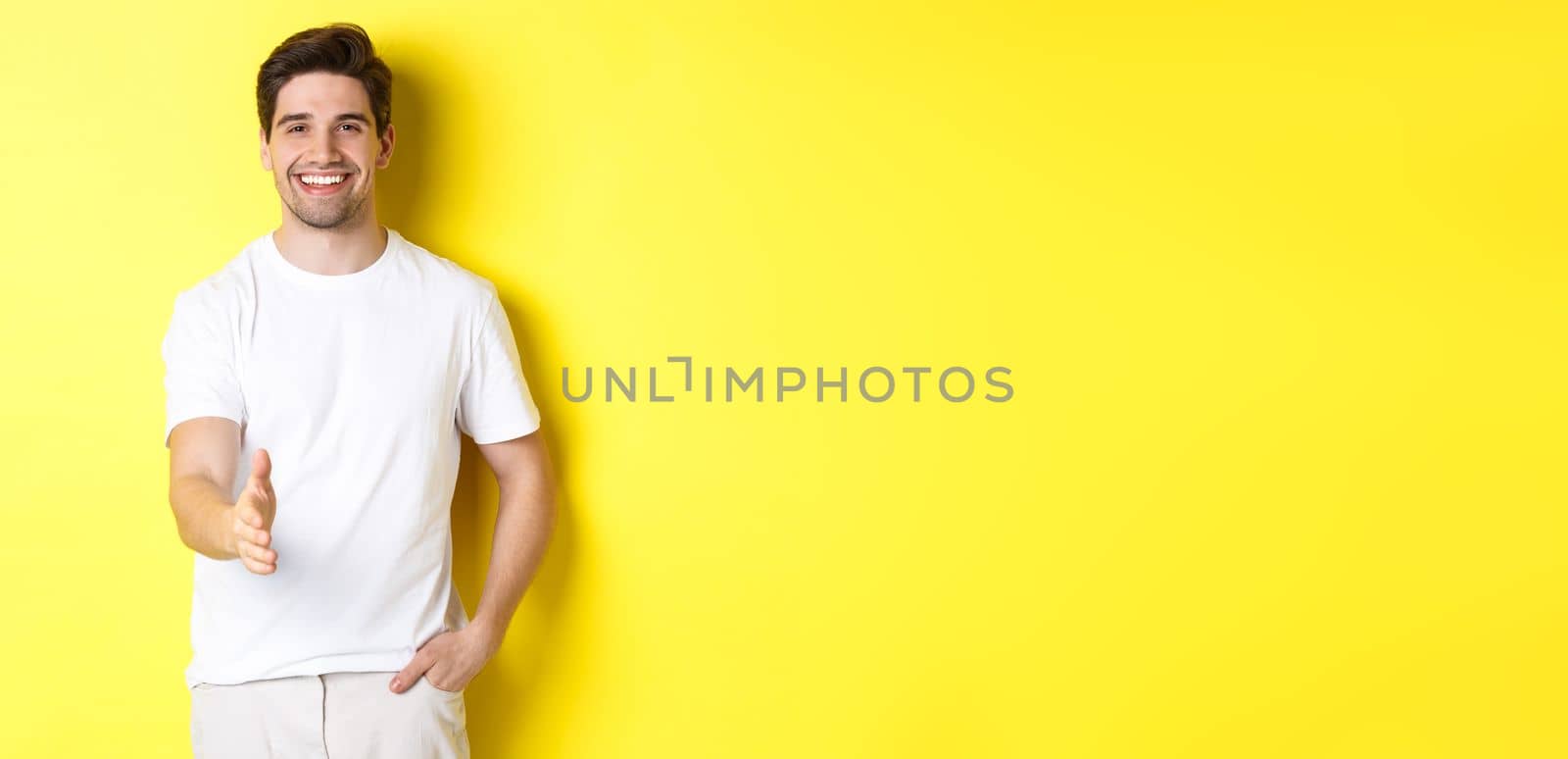 Handsome and confident man extending hand for handshake, greeting you, saying hello, standing in white t-shirt over yellow background by Benzoix