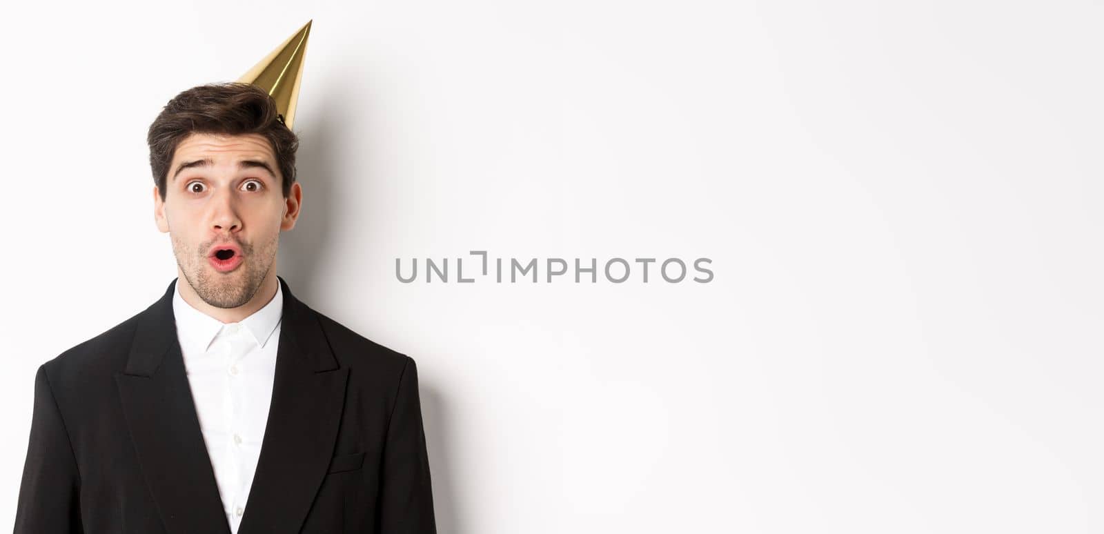 Close-up of attractive man in party hat and trendy suit, looking surprised, celebrating new year, standing against white background.