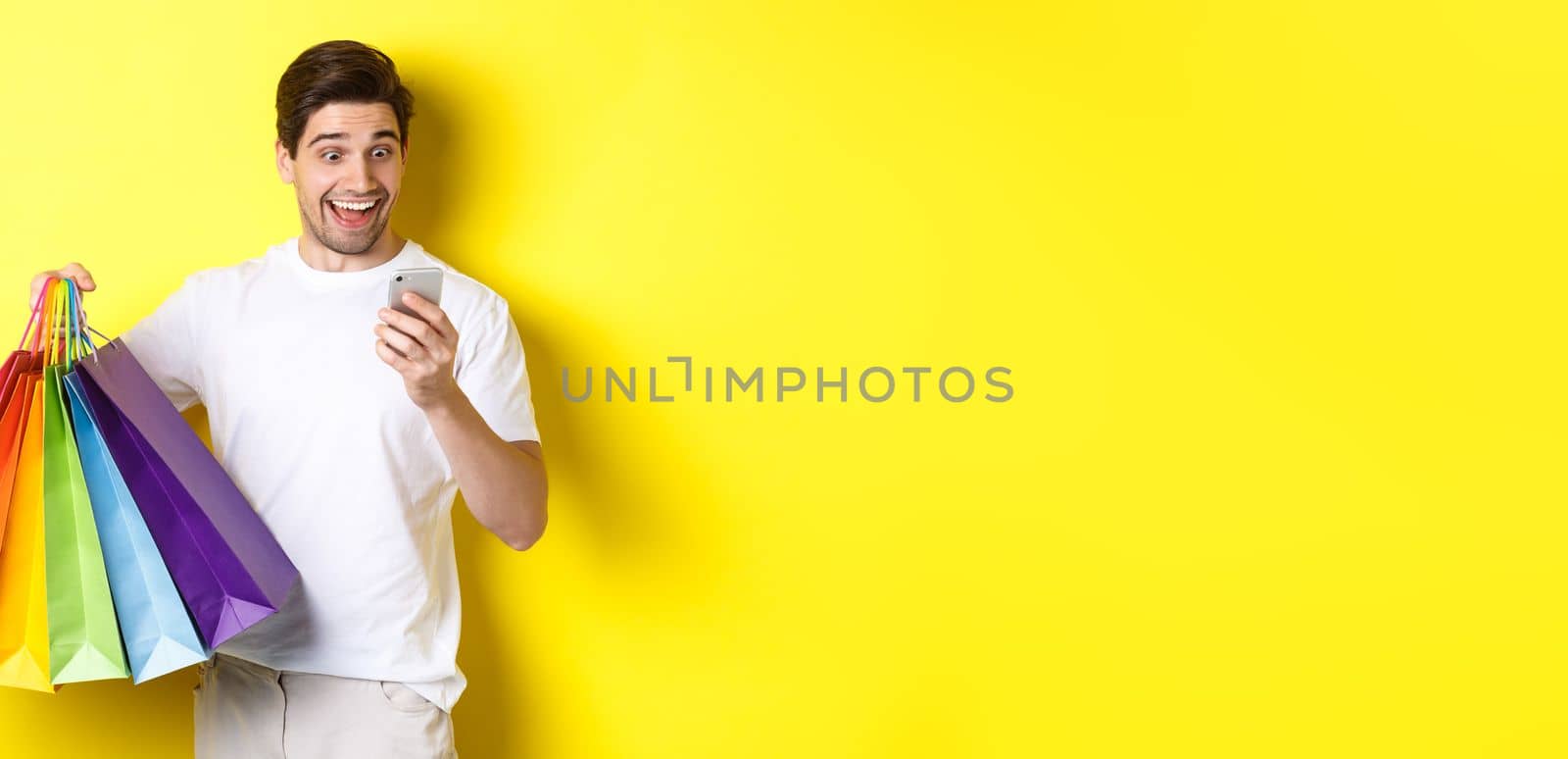 Excited man holding shopping bags and looking happy at mobile phone screen, standing over yellow background by Benzoix