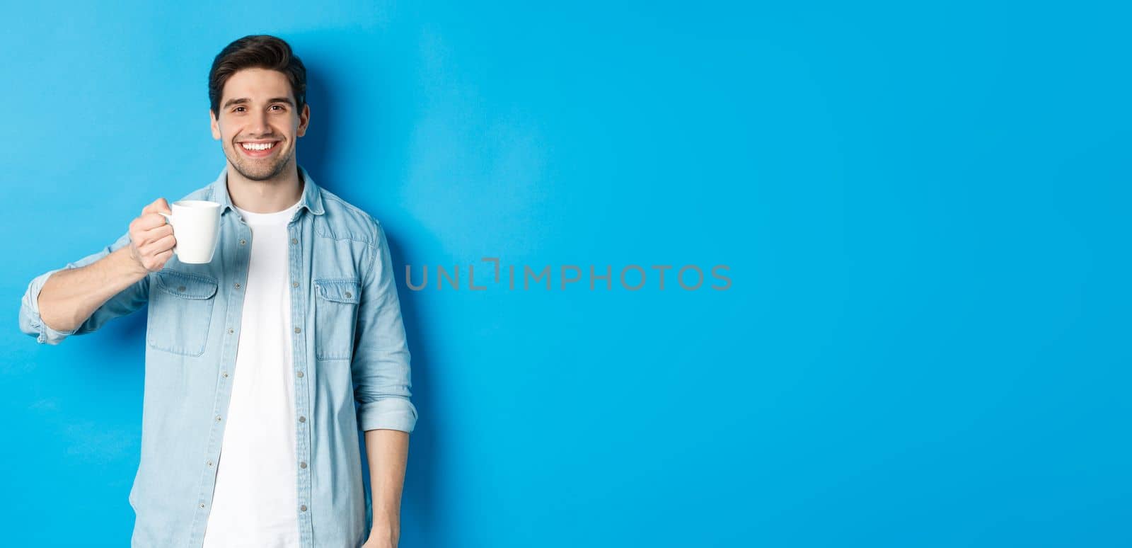 Smiling bearded man holding mug and drinking coffee, standing against blue background.