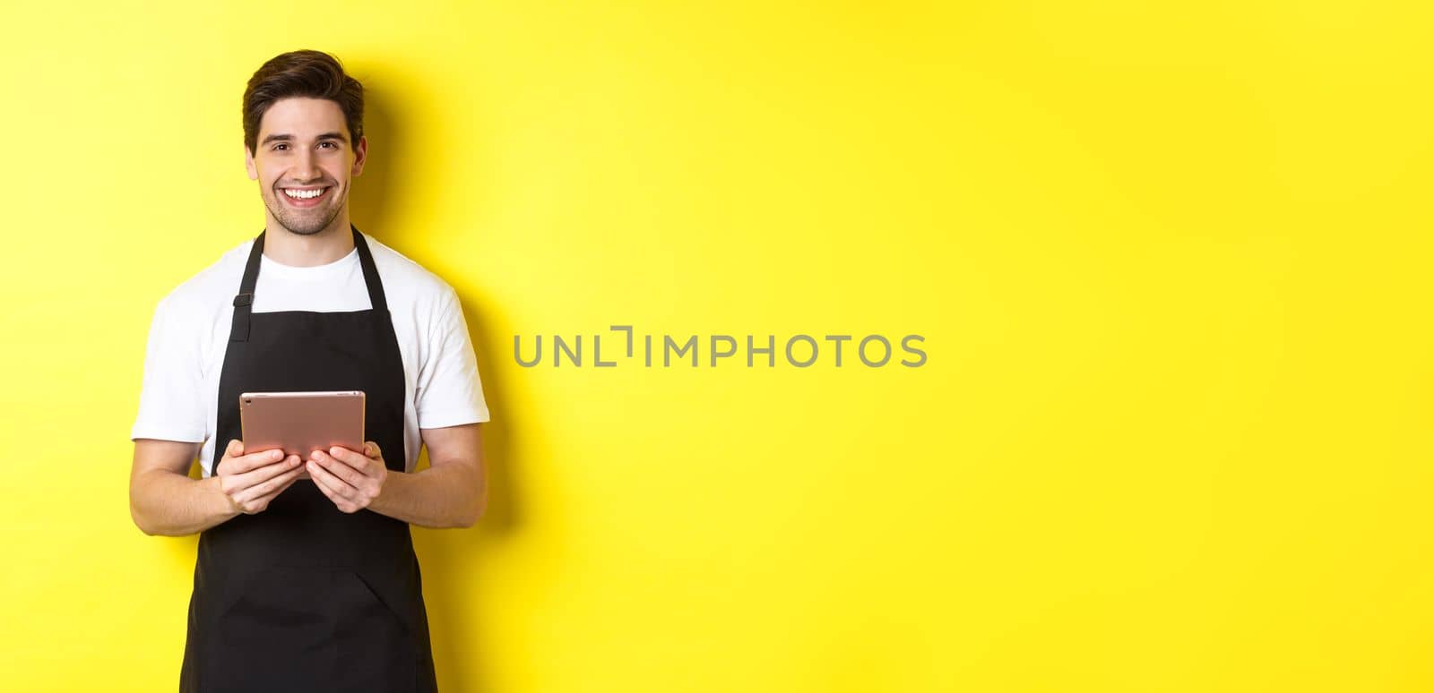 Waiter in black apron taking orders, holding digital tablet and smiling friendly, standing over yellow background.