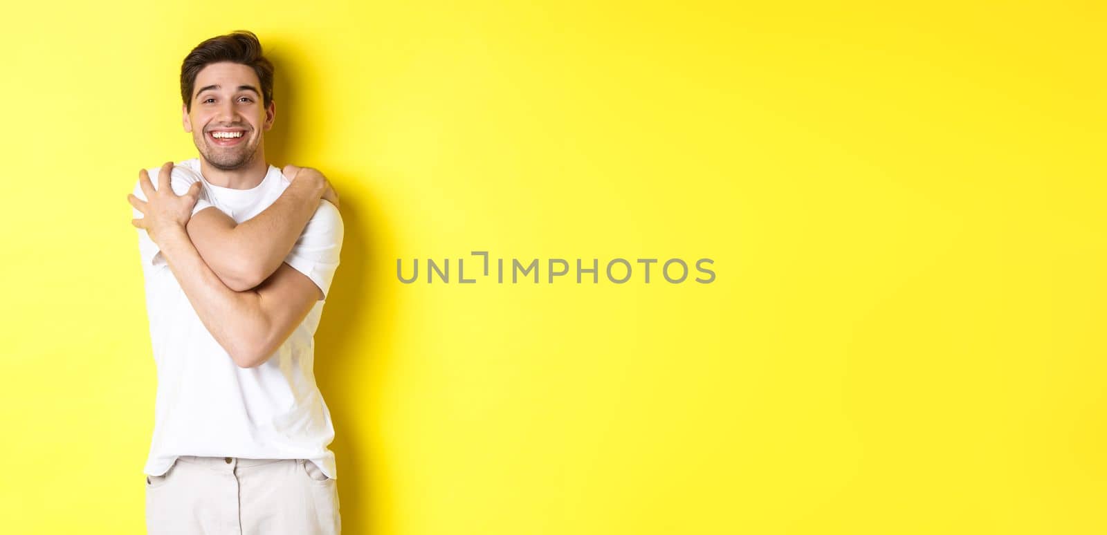 Happy kind guy hugging himself and smiling, standing pleased against yellow background. Copy space