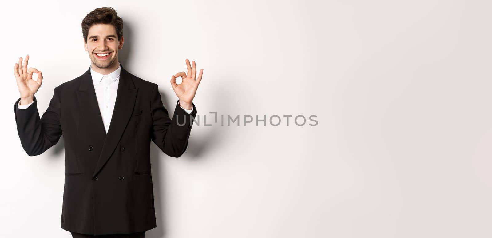 Concept of new year party, celebration and lifestyle. Portrait of handsome attractive man in black suit, smiling and showing okay signs, approve and recommend something, white background.