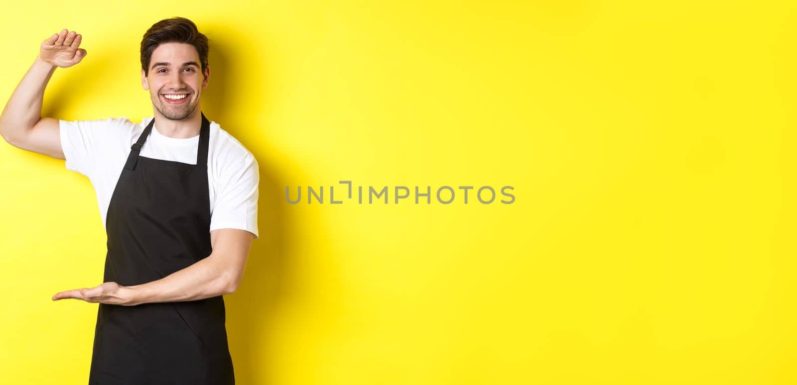 Smiling handsome barista showing something long or large, standing over yellow background.