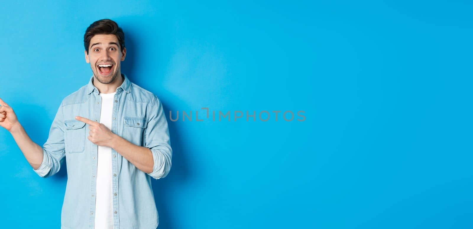 Excited handsome 25s years man with beard, pointing fingers left and smiling amazed, standing against blue background.