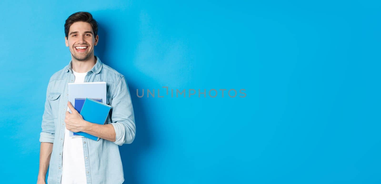 Smiling man studying, holding notebooks and looking happy, standing over blue background.