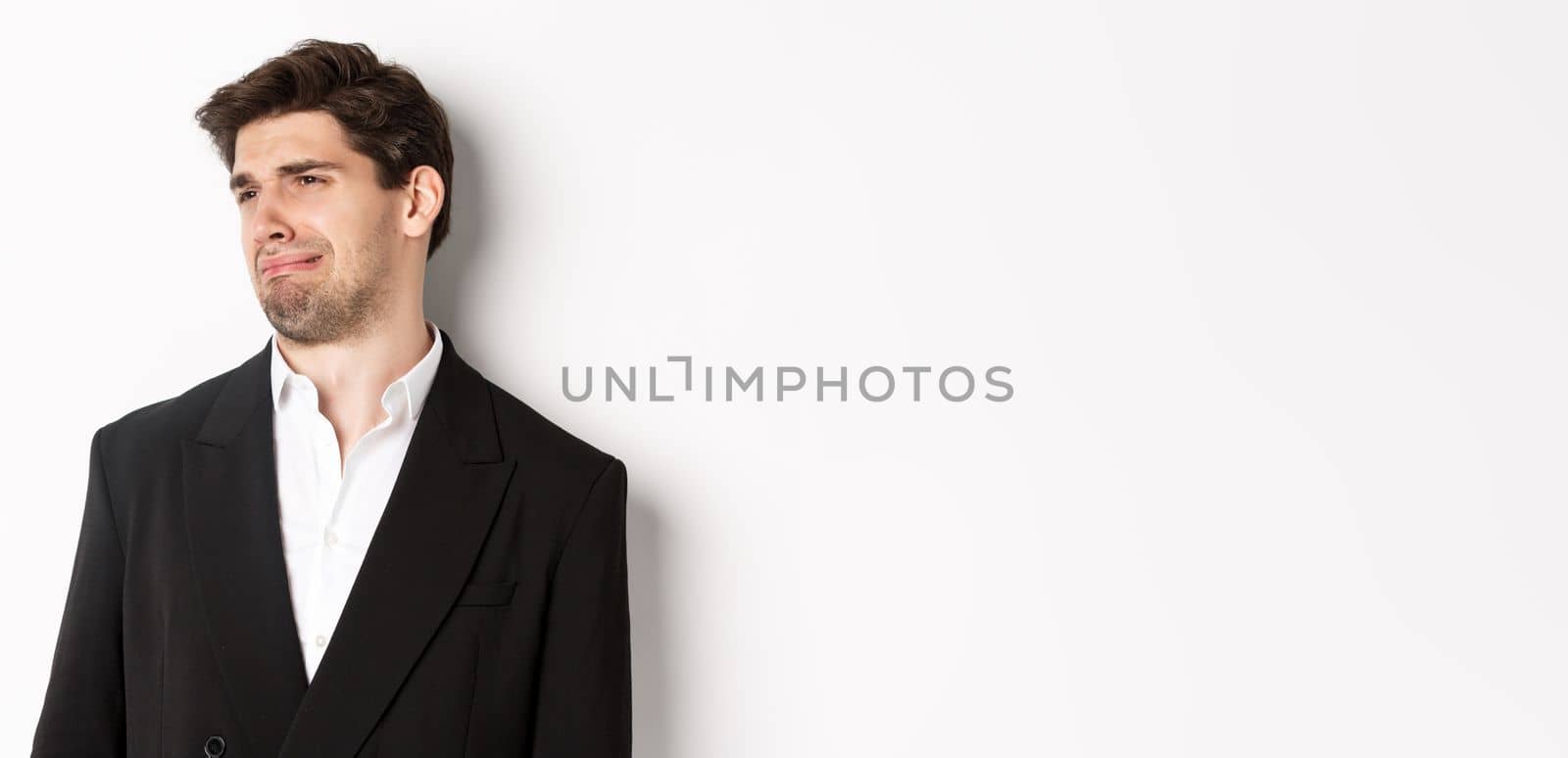 Close-up of disgusted young man in trendy suit, grimacing upset, looking left and standing against white background.