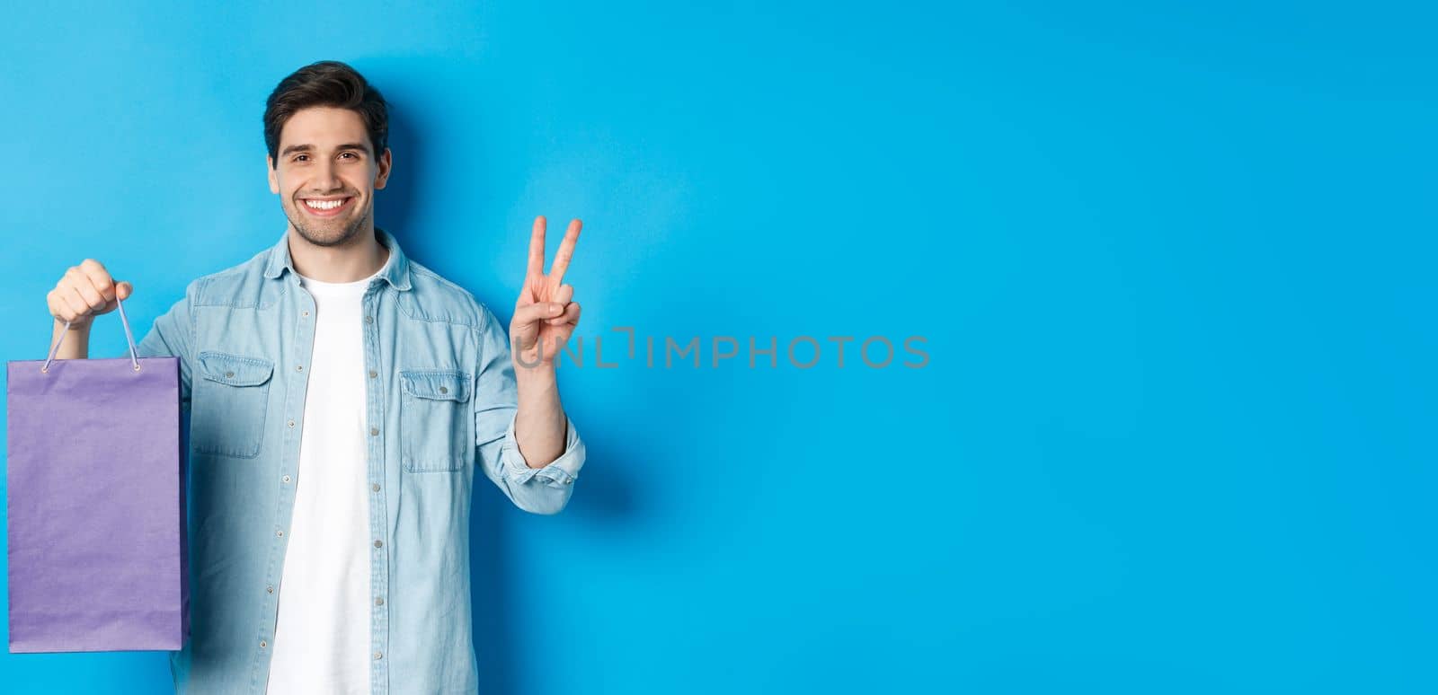 Concept of shopping, holidays and lifestyle. Happy bearded guy holding paper bag from store and showing peace sign, standing over blue background.