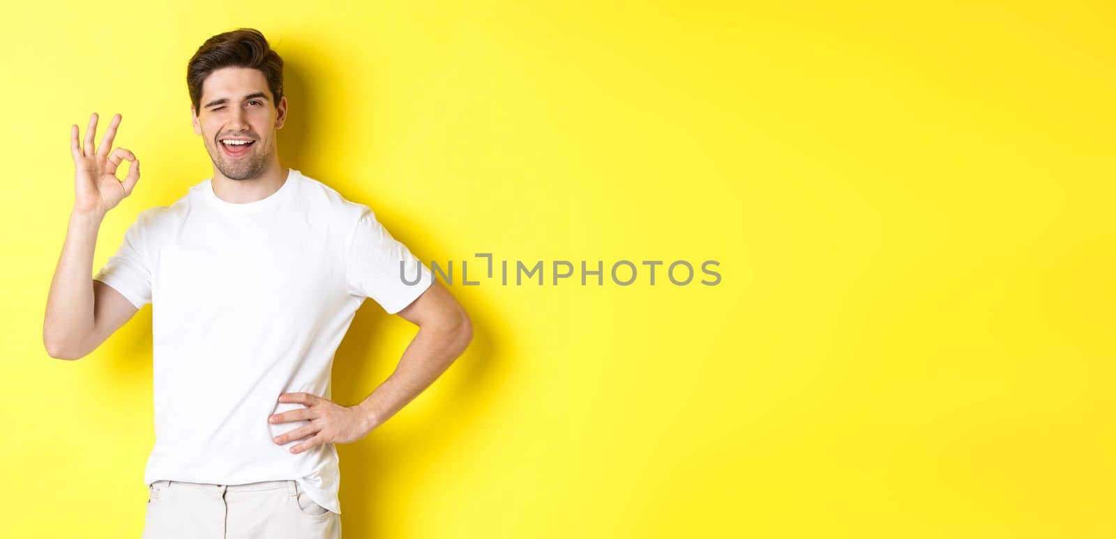 Confident handsome man winking, showing okay sign in approval, like something good, standing over yellow background.