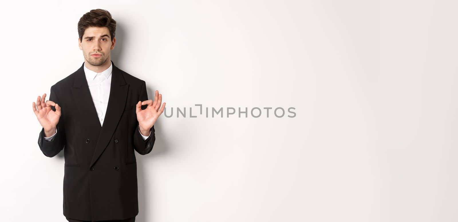 Concept of new year party, celebration and lifestyle. Portrait of confident good-looking man in black suit, showing okay sign and approve something, standing over white background.