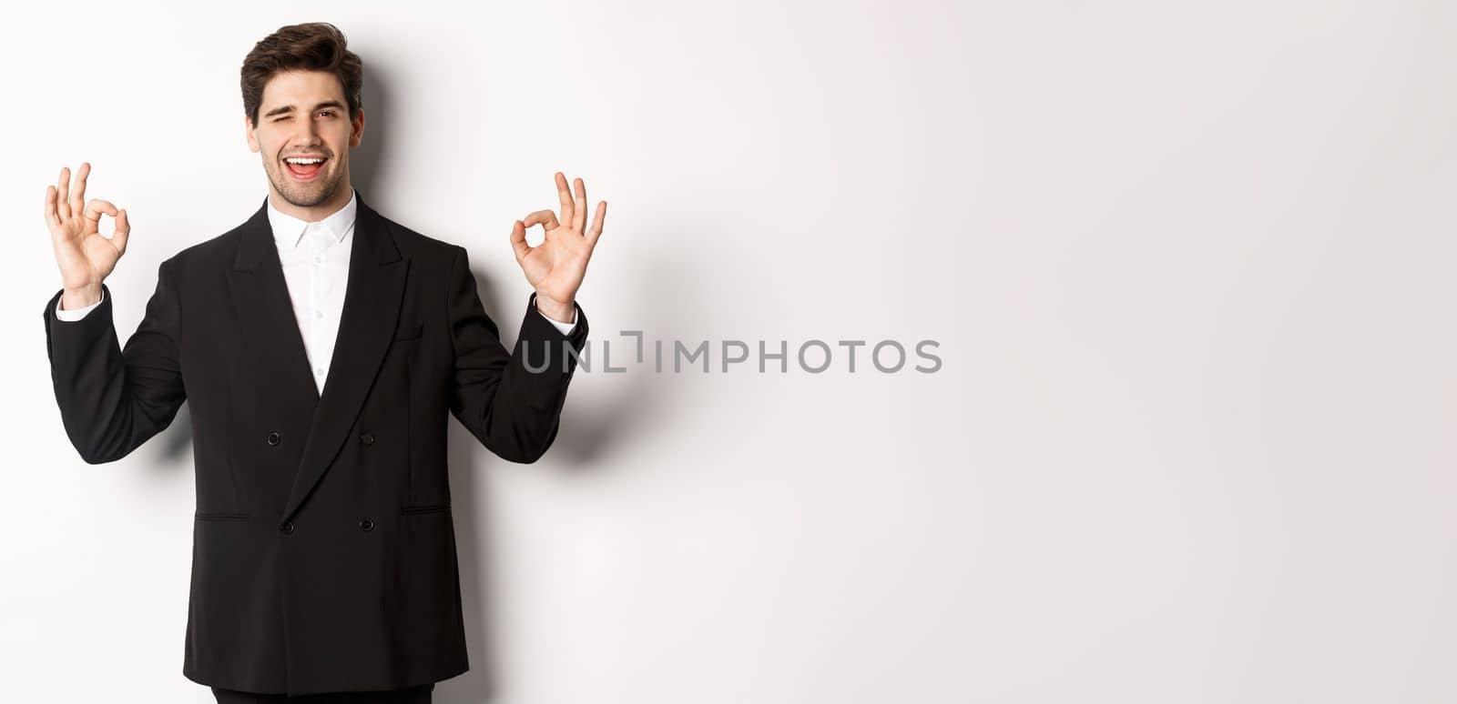 Concept of new year party, celebration and lifestyle. Portrait of successful handsome businessman in suit, winking and showing okay signs, recommending something good, white background.