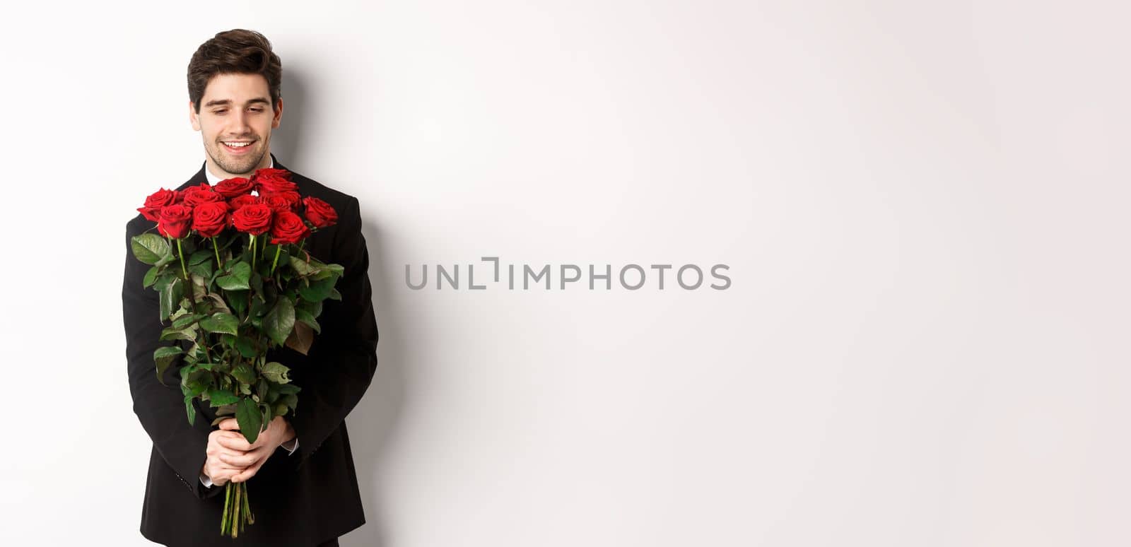 Image of handsome boyfriend in black suit, holding bouquet of red roses and smiling, being on a date, standing over white background by Benzoix