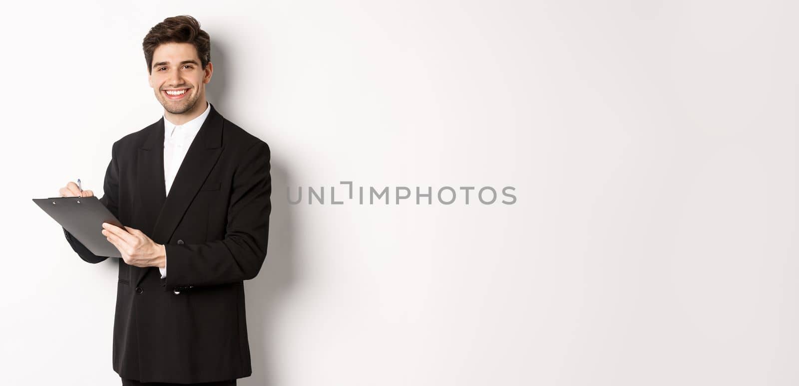 Portrait of confident businessman in black suit, signing documents and smiling, standing happy against white background.