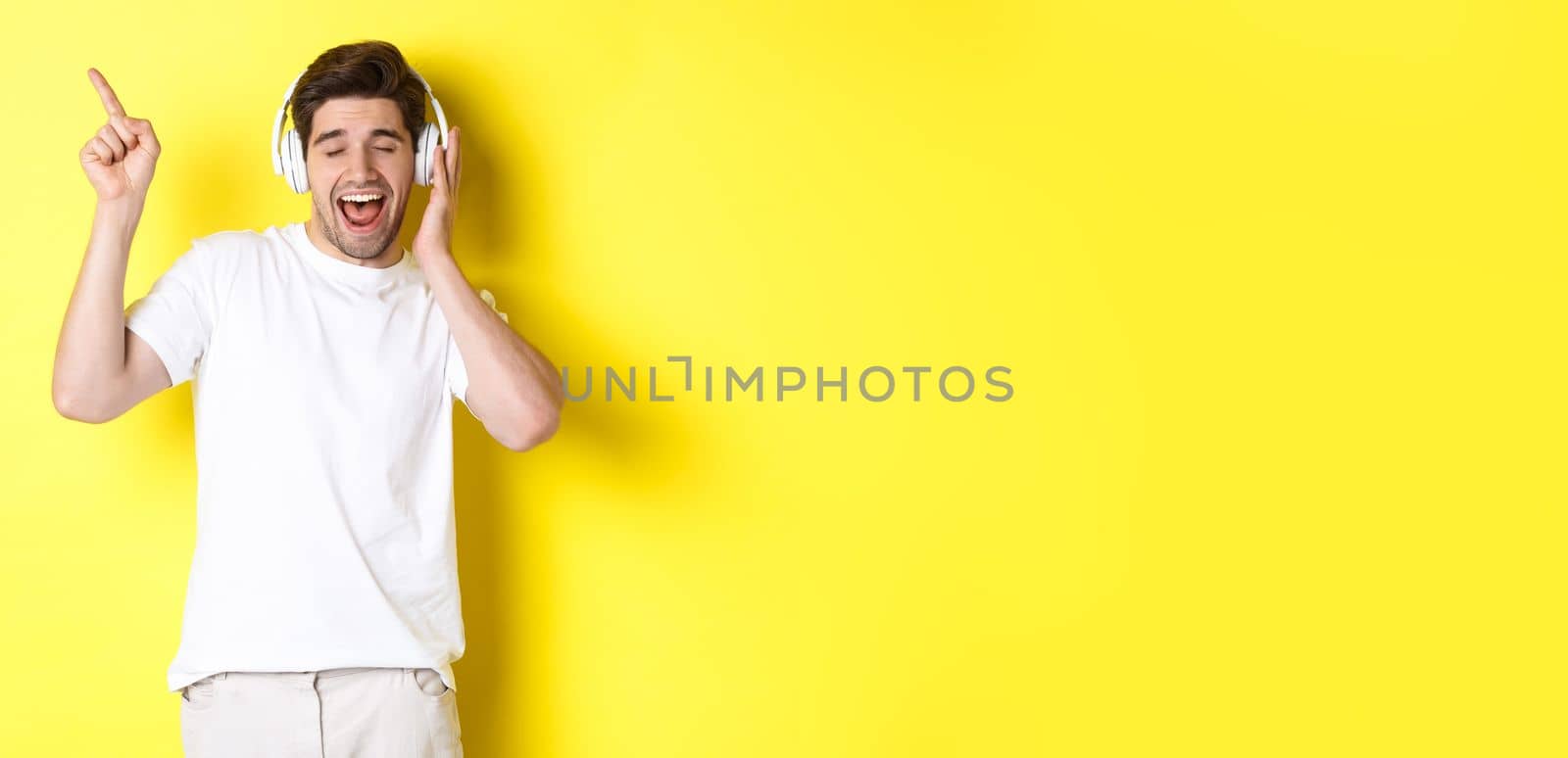 Happy man listening music in headphones, pointing finger at promo offer for black friday, standing over yellow background.
