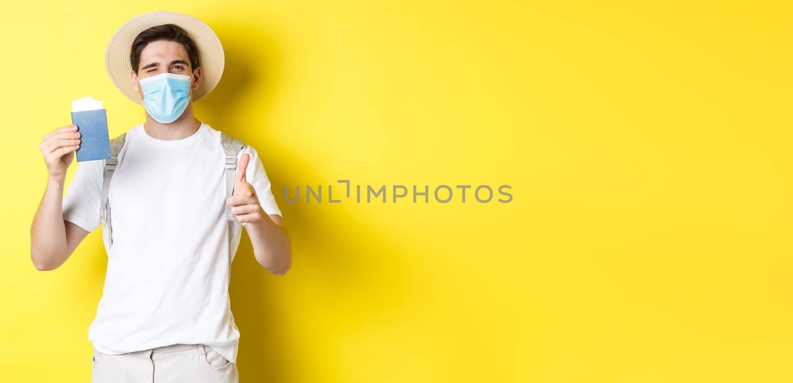 Concept of covid-19, tourism and pandemic. Happy guy tourist in medical mask showing passport with tickets for vacation, winking and pointing at camera, yellow background by Benzoix