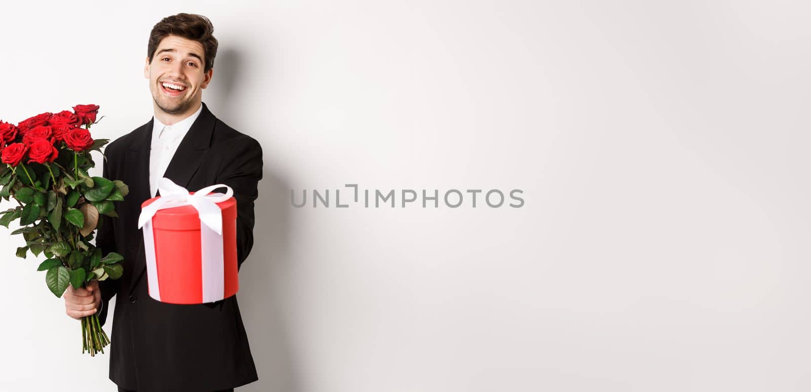 Concept of holidays, relationship and celebration. Image of handsome smiling guy in black suit, holding bouquet of red roses and giving you a gift, standing against white background by Benzoix